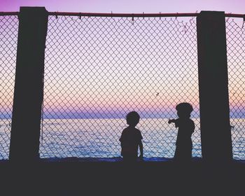 Silhouette kids by chainlink fence at beach during sunset