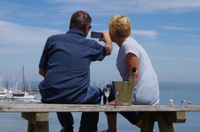 Rear view of couple taking selfie while sitting on bench against lake