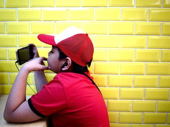 Boy watching mobile phone while sitting at table by yellow wall
