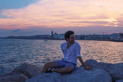 Woman sitting on rock at shore against sky during sunset