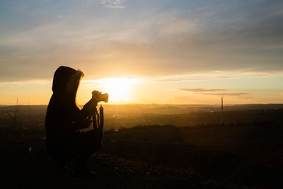 Silhouette woman holding camera against sky during sunset