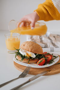 Breakfast sandwich with egg and arugula and cherry tomatoes on the table, woman pours orange juice