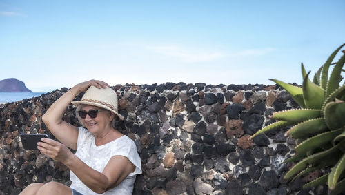 Smiling senior woman taking selfie while sitting by retaining wall