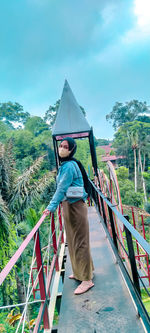 Low angle view of young woman standing on footbridge against sky