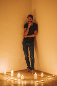 Thoughtful man standing by lit candles against wall in darkroom at home