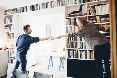 Rear view of man giving book to woman standing against bookshelf at home