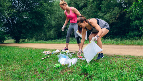 Full length of happy women picking up garbage while crouching on grass against trees