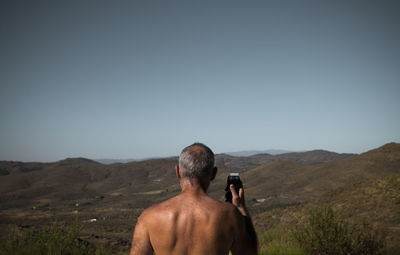 Rear view of shirtless adult man shaving himself with shaver against mountain and sky