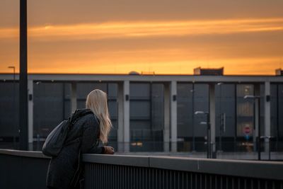 Side view of woman standing by railing in city against sky during sunset