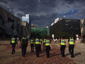 Rear view of policemen walking at federation square against cloudy sky at dusk