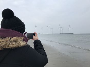 Rear view of women standing on beach against sky taking picture with mobile