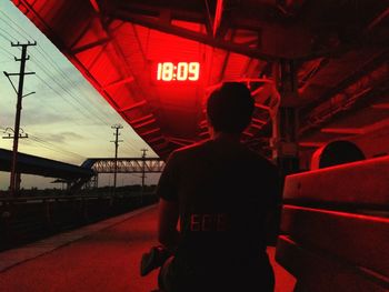 Rear view of man sitting at illuminated railroad station platform during dusk