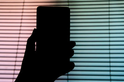 Cropped hand holding mobile phone against window blinds