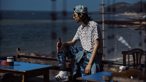 Young man holding beer bottle while looking at sea