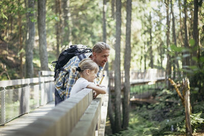 Smiling father with backpack talking to daughter on footbridge in forest