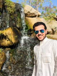 Portrait of young man wearing sunglasses standing against waterfall