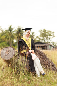 Young woman in graduation gown sitting on tree stump at field