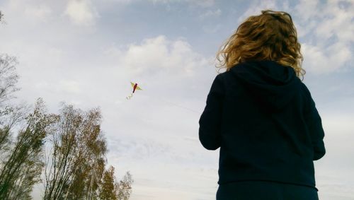 Rear view of woman flying kite while standing against cloudy sky