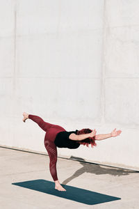 Side view of barefooted woman in sportswear doing yoga in warrior three pose on mat training alone on street against concrete wall in sunlight