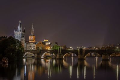 Illuminated bridge over river by buildings against sky at night