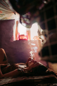 Side view of resting woman in casual wear lying on bed and smoking blunt at home