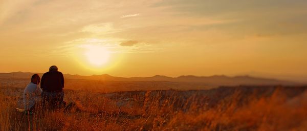 Father and son sitting on field against sky during sunset