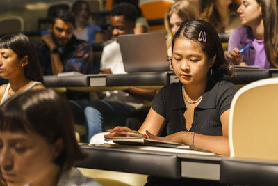 Young female student reading book while sitting in lecture hall at university
