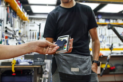 Low angle of crop client using smartphone to pay to male technician in repair workshop