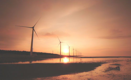Silhouette wind turbines on shore against sky during sunset