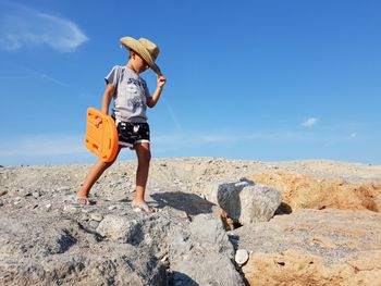 Full length of boy wearing hat standing on rock against blue sky during sunny day