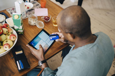 High angle view of man shopping online on digital tablet through credit card in dining room at home