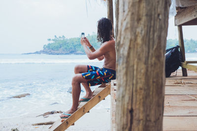 Full length of shirtless man using mobile phone while sitting on wood