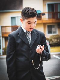 Boy in suit holding pocket watch while standing in city