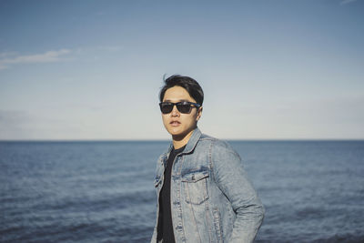 Young man wearing sunglasses and denim jacket while standing against sea and sky