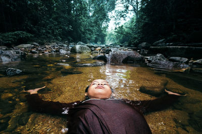 Boy lying in stream amidst rocks in forest