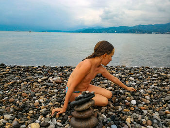 Shirtless girl stacking stones at beach against sky