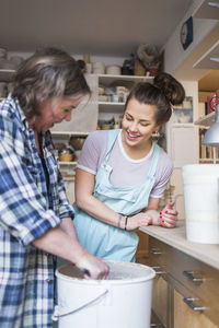Happy woman looking at mature female potter mixing clay in bucket at store