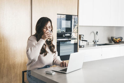 Serious female freelancer sitting at table in kitchen and browsing netbook while working on project remotely while having a coffee