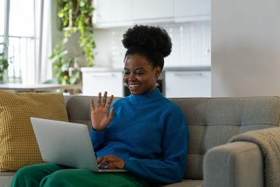 Cheerful young african american woman with smile making video call sits on sofa with laptop on lap