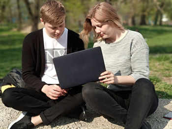 Friends using laptops while sitting in park during sunny day