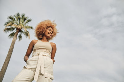 Smiling young woman with hands in pockets standing by palm tree