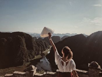 Rear view of woman with arms raised holding conical hat while standing on mountain against sky