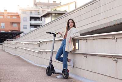 Young woman leaning against the wall holding her e-scooter on the hand in a town park 