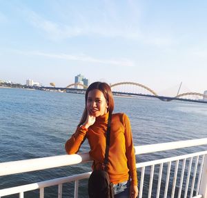 Young woman standing by railing against bridge against sky