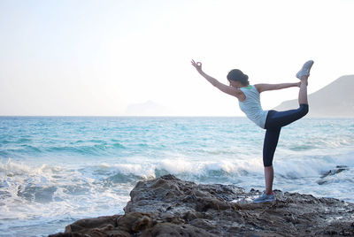 Rear view of woman doing yoga at rocks beach against clear sky 