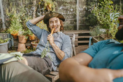 Young male farmer eating carrot sitting by man in urban garden