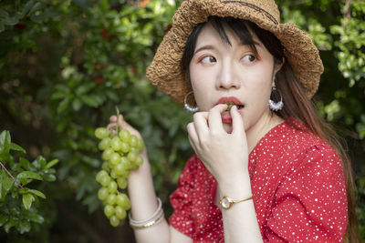 Woman with hat eating grapes while holding bunch by plants
