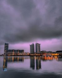 Illuminated buildings by river against sky at dusk