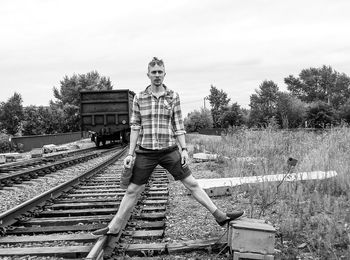 Portrait of man standing on railroad tracks and electric box against sky