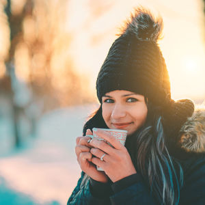 Portrait of young woman holding mug while standing outdoors during sunset